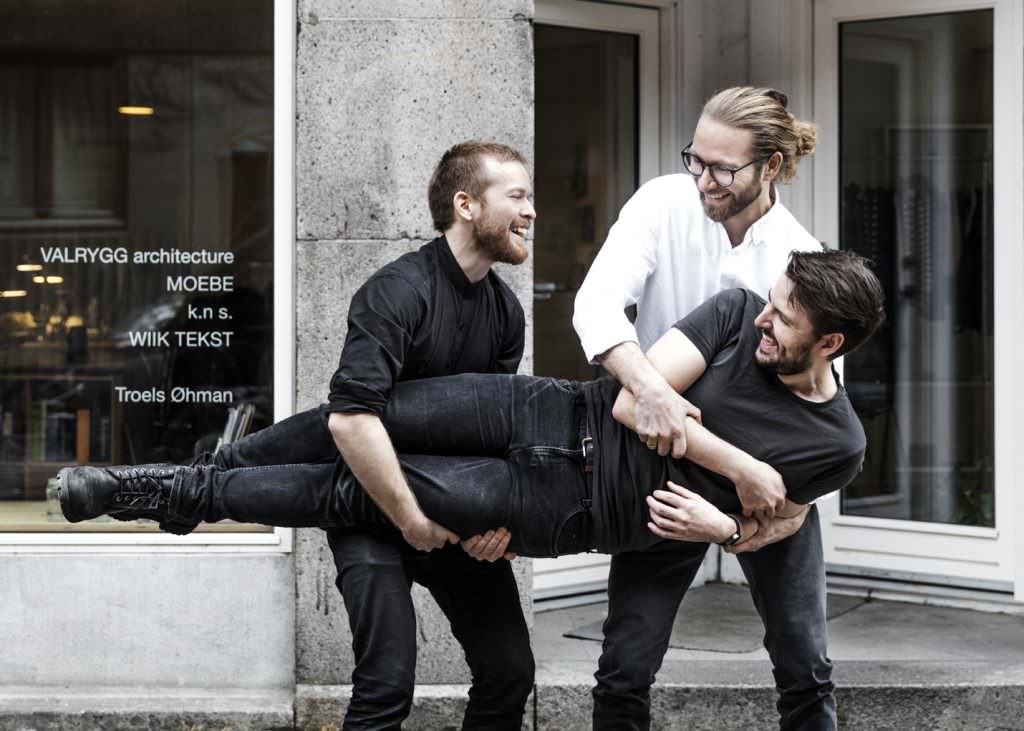 Drei Männer in einem Raum, einer hält den anderen kopfüber, schwarze Shirts, lebhafte Stimmung