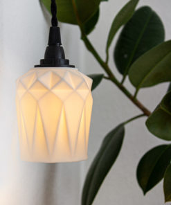 Pendelleuchte aus gefaltetem Porzellan in Creme-Weiß mit schwarzer Textilkordel und natürlichem Pflanzenhintergrund