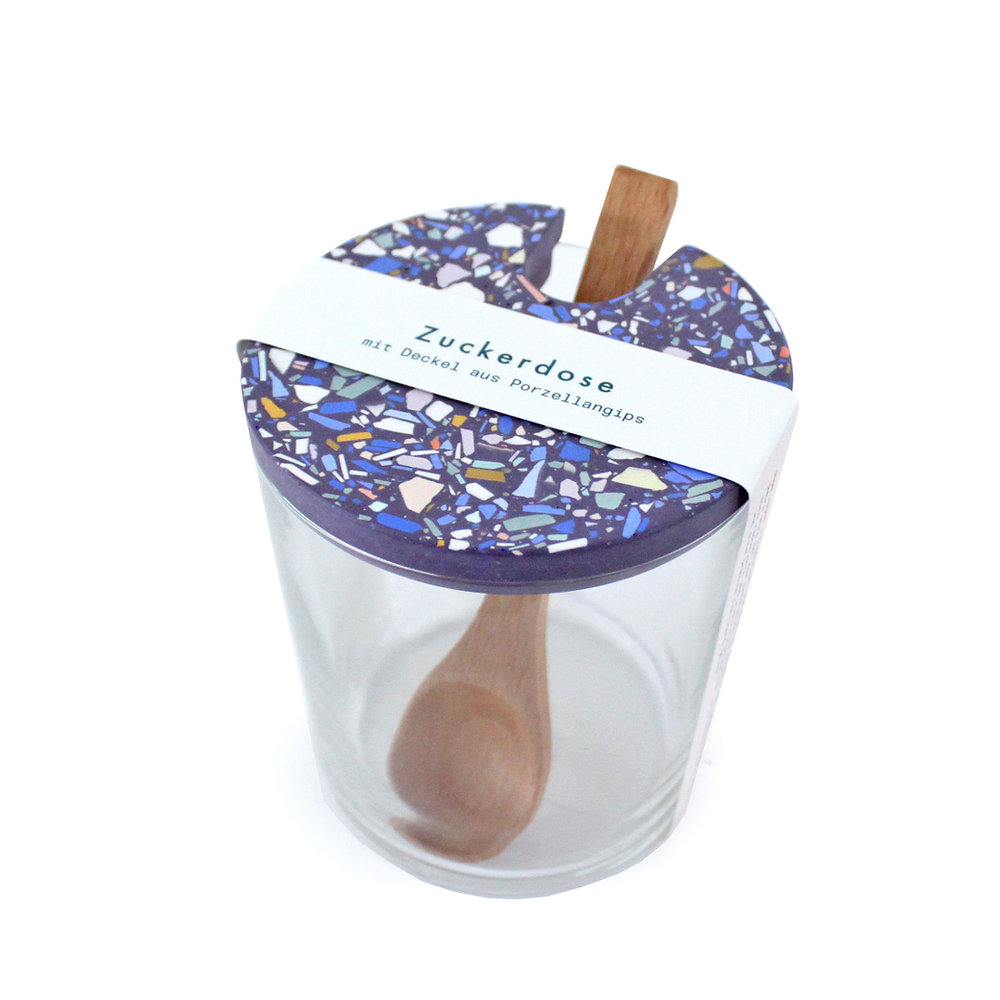 Design-Zuckerdose aus Glas mit violettem Terrazzo-Deckel und Holzlöffel auf weißem Hintergrund