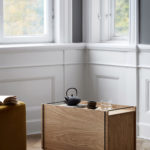 Moderner Holztisch mit Teekanne, Keramiktasse und Wasserglas auf weißem Fenster und raffinierter Wandpaneelierung