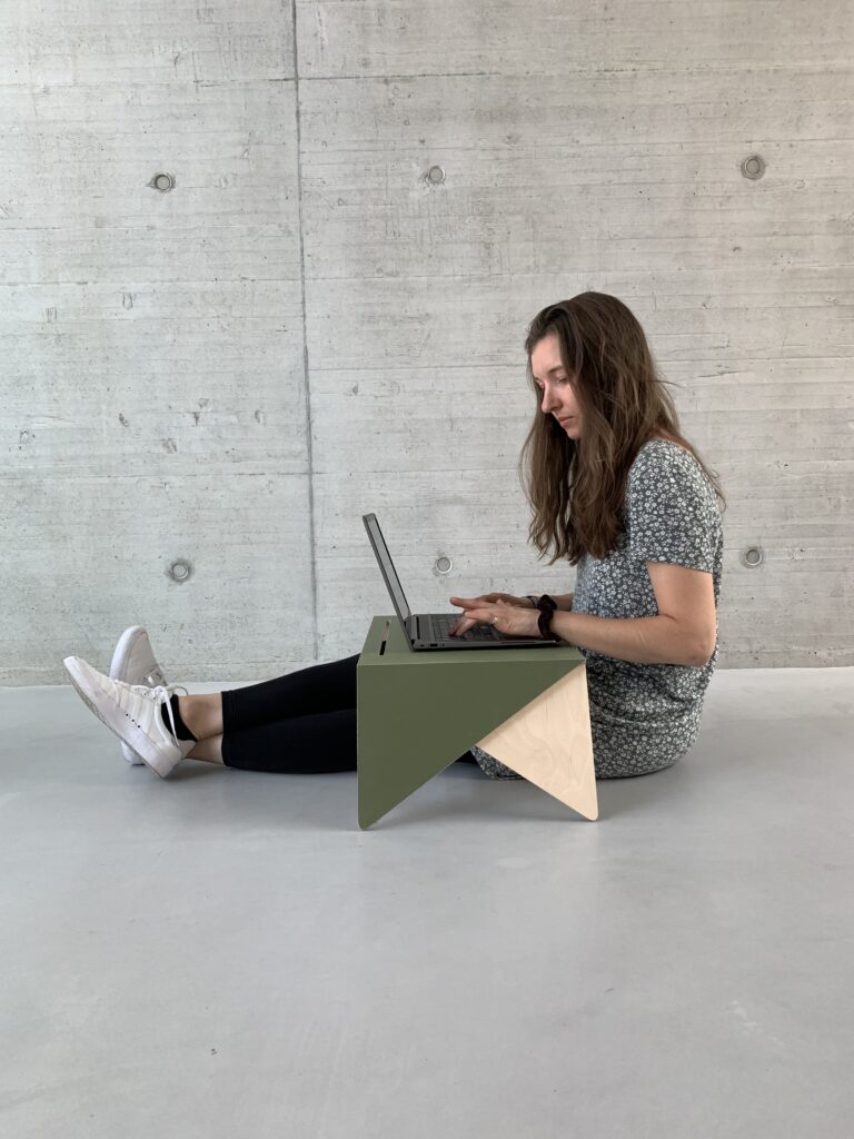 Eine Frau sitzt an einem hölzernen, grünen Tisch mit Laptop. Sie trägt eine schwarze und weiße Bluse