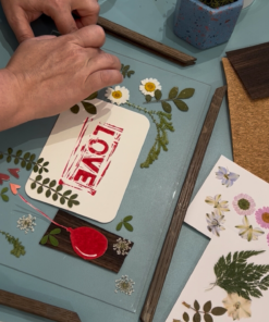 Person gestaltet 'LOVE'-Motiv mit Blumen und Blättern auf Holzplatte im Workshop